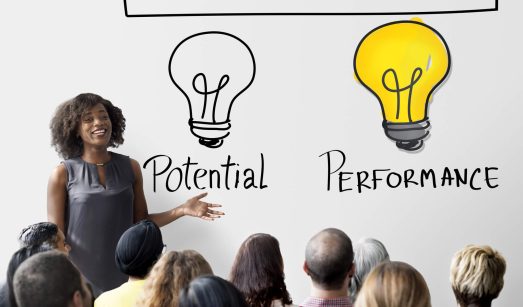 תמונת נושא של המאמר קורס אימון אישי המציגה אישה מחייכת מציגה מול קהל על קיר לבן כתוב Let Yours Potential Shine לצד ציור של נורה כבויה המסומנת Potential ונורה דולקת המסומנת Performance.